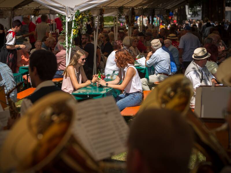 Beim Klosterfest sitzen zwei Frauen gemeinsam unter einem Festzelt auf einer Bierbank. Sie sind einander zugewandt und unterhalten sich. Im Hintergrund sind weitere Menschen und festliche Dekorationen zu sehen.