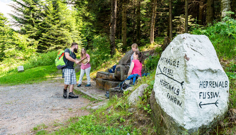 Vier Wanderer die Ihre Wasserflaschen an einem Brunnen auffüllen und rechts im Bild ein Stein ist, dr mit weiße Farbe besprüht wurde und Wegweiser aufgemalt sind.