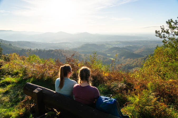 Zwei Personen sitzen im Vordergrund auf einer Bank und betrachten den Ausblick in die Landschaft