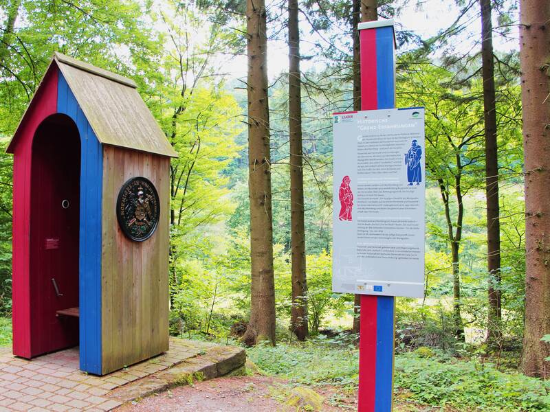 links ein Häuschen in blau, rot gestrichen, rechts ein Erklärschild