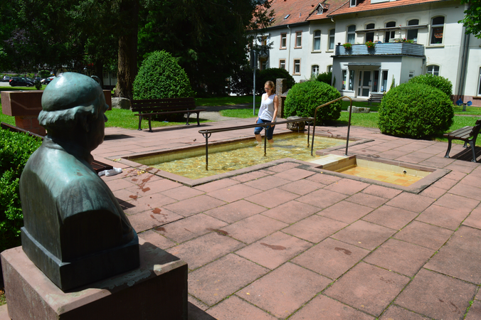 Im Vordergrund ist einen Statue zusehen, im Hintergrund geht eine Frau durch ein Wassertretbecken+