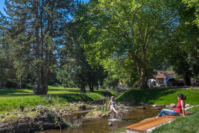 Im Fluss steht eine Frau die mit Wasser spritzt und auf einer Parkbank sitzt eine Frau die ihr zusieht#