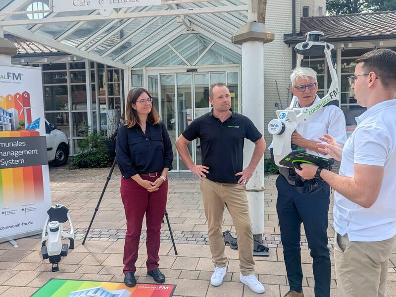 Vier Personen stehen vor einem Gebäude und diskutieren, während ein Plakat mit der Aufschrift "Kommunales Energiemanagement mit System" im Vordergrund zu sehen ist.
