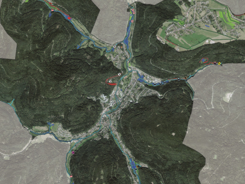 Das Bild zeigt eine Satellitenansicht eines bergigen Gebietes mit einem zentral durchfließenden Fluss und mehreren Straßen, die sich durch bewaldete Hügel und eine kleine Ortschaft schlängeln.