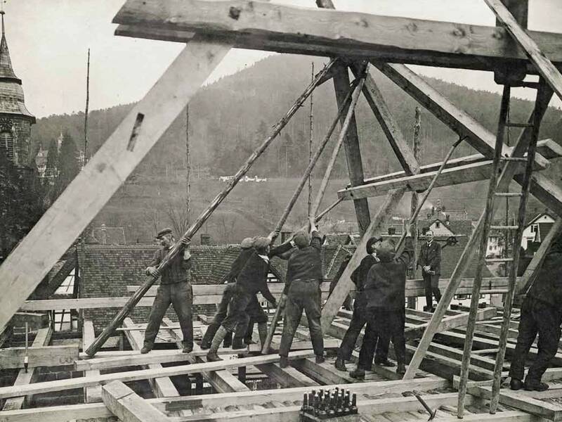 Mehrere Arbeiter errichten ein Dachgerüst aus Holz auf einer Baustelle mit einem Kirchturm und bewaldeten Hügeln im Hintergrund.