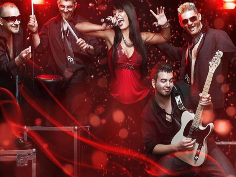 Eine Band aus fünf Mitgliedern, darunter eine Sängerin in rotem Outfit und Musiker mit Instrumenten, tritt energetisch auf einer Bühne mit roten Lichteffekten auf.