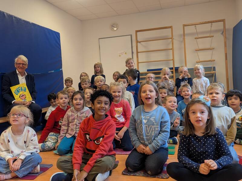 Ein Mann sitzt mit einem Buch in der Hand neben einer großen Gruppe von lächelnden Kindern in einem Raum mit Sportgeräten.