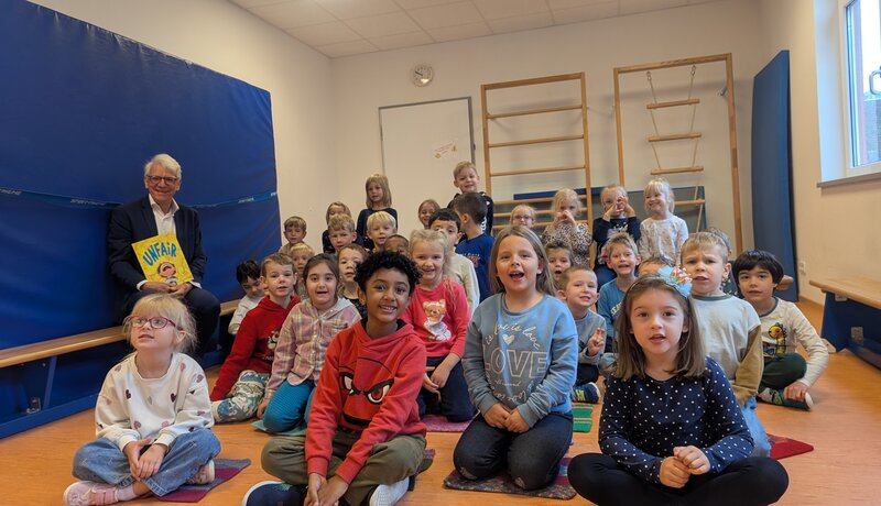 Ein Mann sitzt mit einem Buch in der Hand neben einer großen Gruppe von lächelnden Kindern in einem Raum mit Sportgeräten.