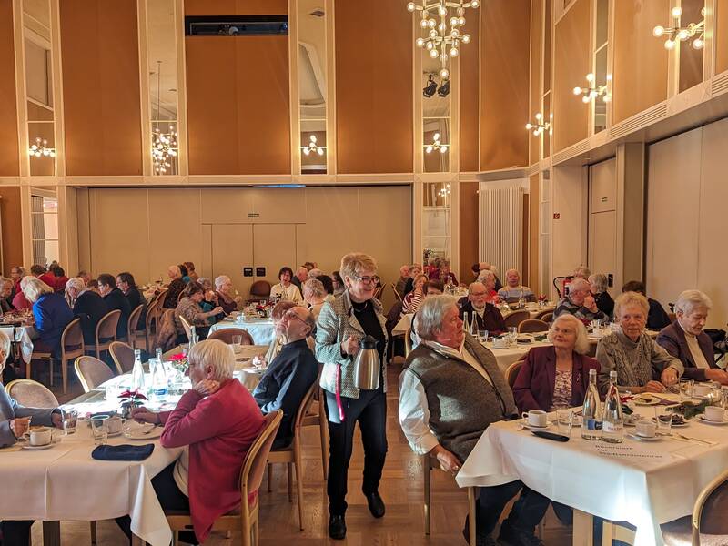 In einem großen, elegant beleuchteten Raum sitzen viele ältere Menschen an runden, gedeckten Tischen und unterhalten sich bei Getränken und Speisen.