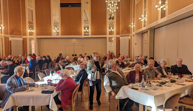 In einem großen, elegant beleuchteten Raum sitzen viele ältere Menschen an runden, gedeckten Tischen und unterhalten sich bei Getränken und Speisen.