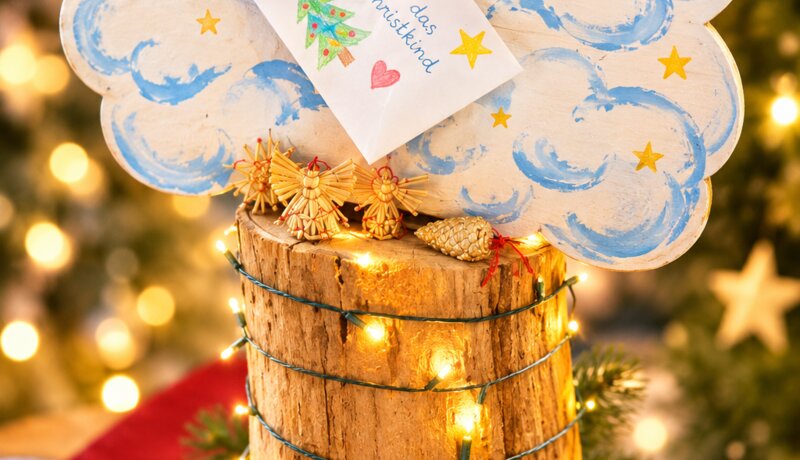 Ein dekorativer "Himmelsbriefkasten" in Form einer Wolke steht auf einem mit Lichtern umwickelten Holzstamm, umgeben von weihnachtlichen Ornamenten und Tannenzapfen; darin steckt ein Brief "An das Christkind".