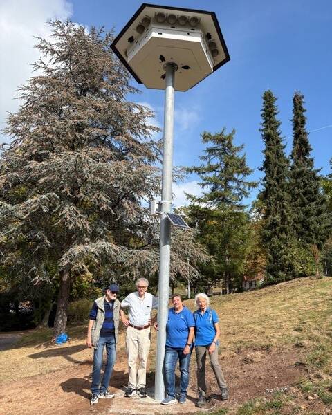 Vier Personen stehen neben einem hohen Mast mit einem großen, sechseckigen Vogelhaus und Solarpanel, umgeben von Bäumen und Wiese.