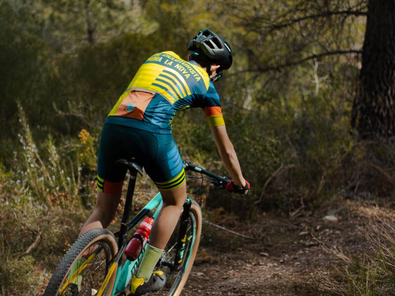 Eine Person in buntem Radsportoutfit fährt ein Mountainbike auf einem schmalen Waldweg.