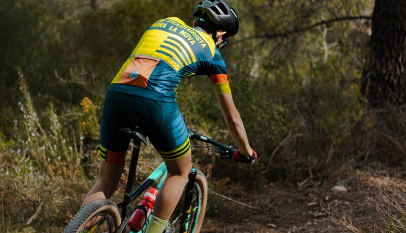 Eine Person in buntem Radsportoutfit fährt ein Mountainbike auf einem schmalen Waldweg. | © Pexels/Marius Mitreanu