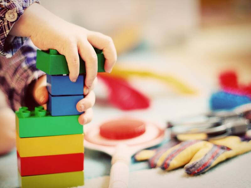 Ein Kind stapelt bunte LEGO-Steine, umgeben von verschiedenen Spielzeugen auf dem Boden.